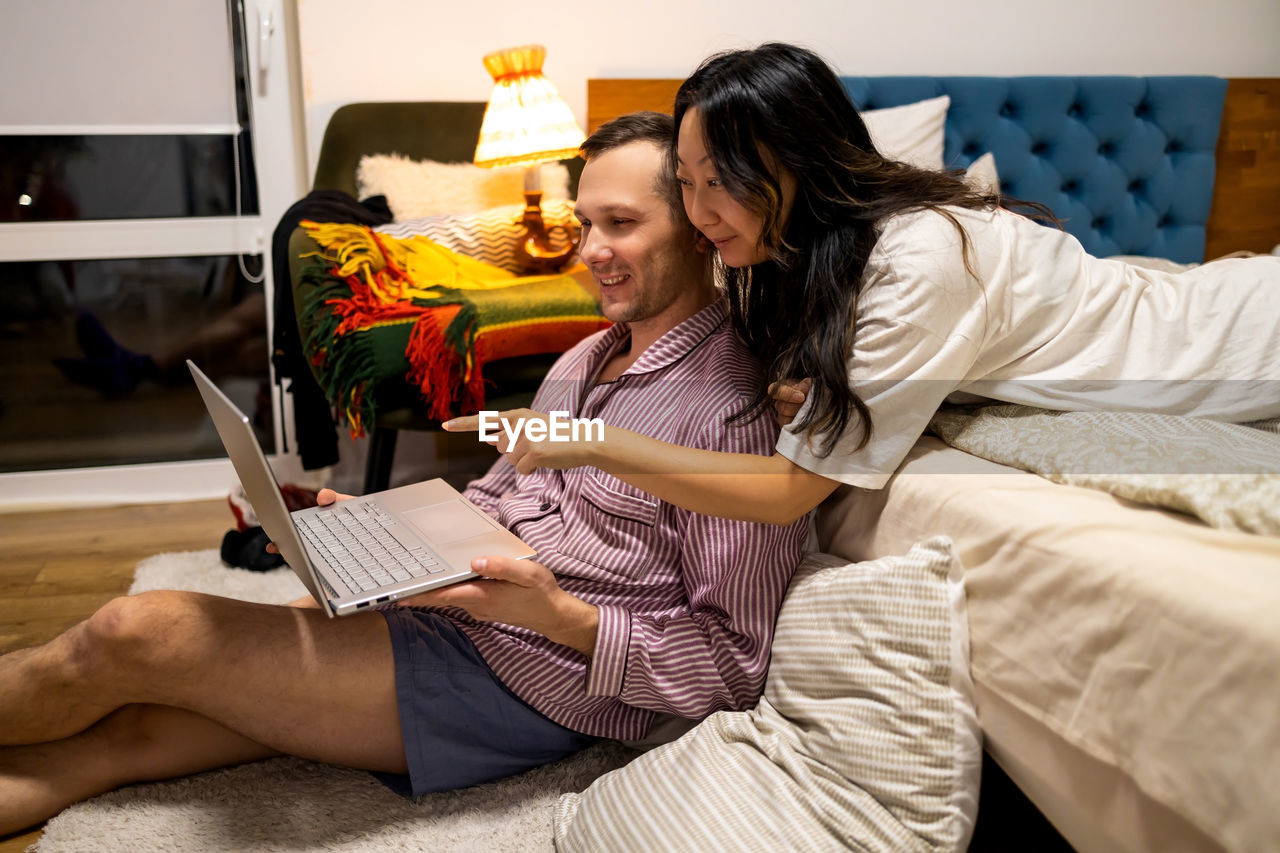 Couple watching funny content on laptop while resting on bed at home in evening.