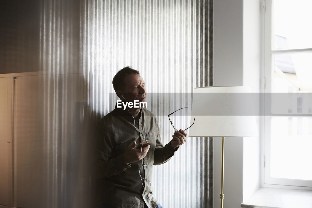 Mature businessman holding eyeglasses while talking on smart phone through headphones standing against wall at creative