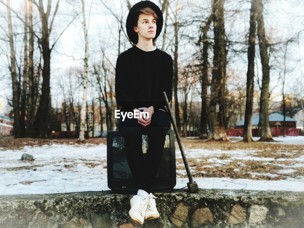 Young man looking away while sitting on television set in forest during winter