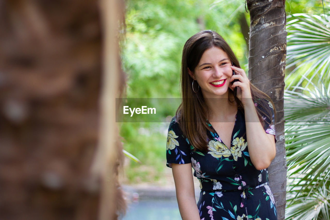 Beautiful young woman using phone while standing against tree trunk