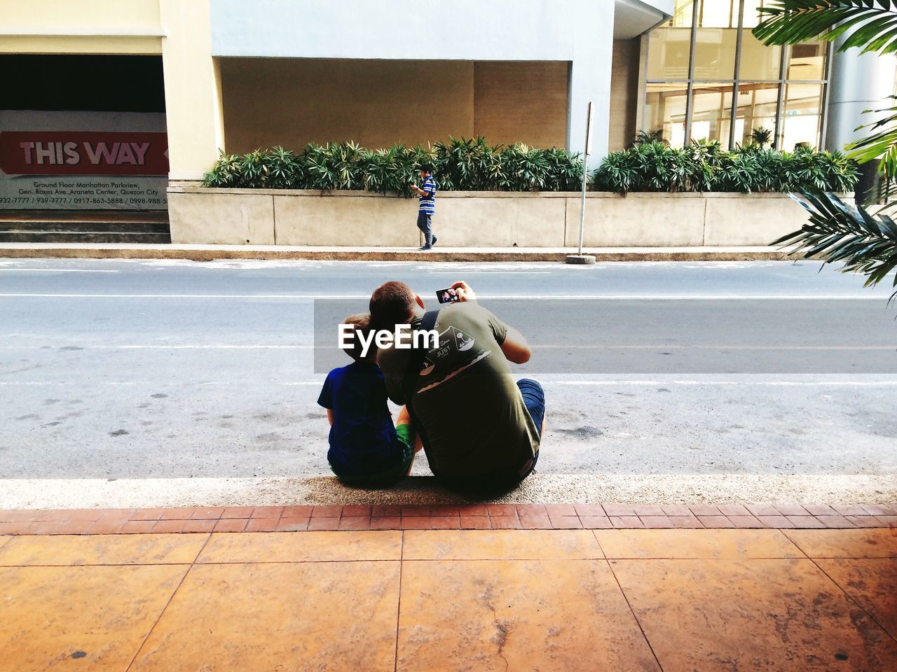 Rear view of father and son photographing while sitting on footpath by street