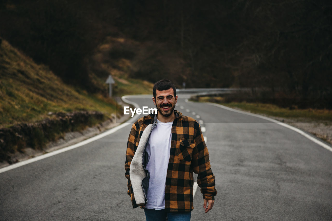 Bearded black haired cheerful man in casual wear smiling and looking at camera while standing on empty road in autumn countryside