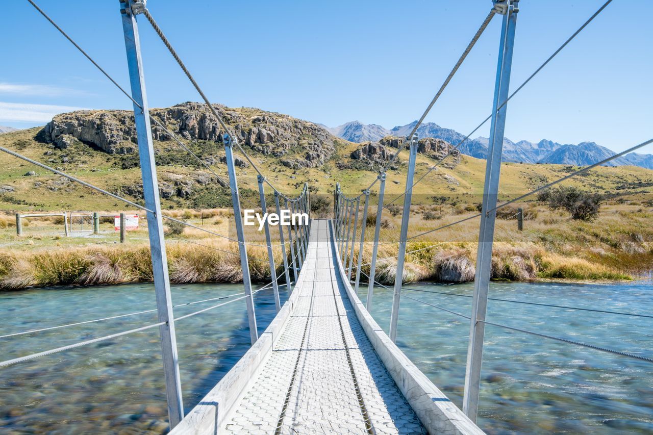 Empty footbridge over river against mountains and blue sky