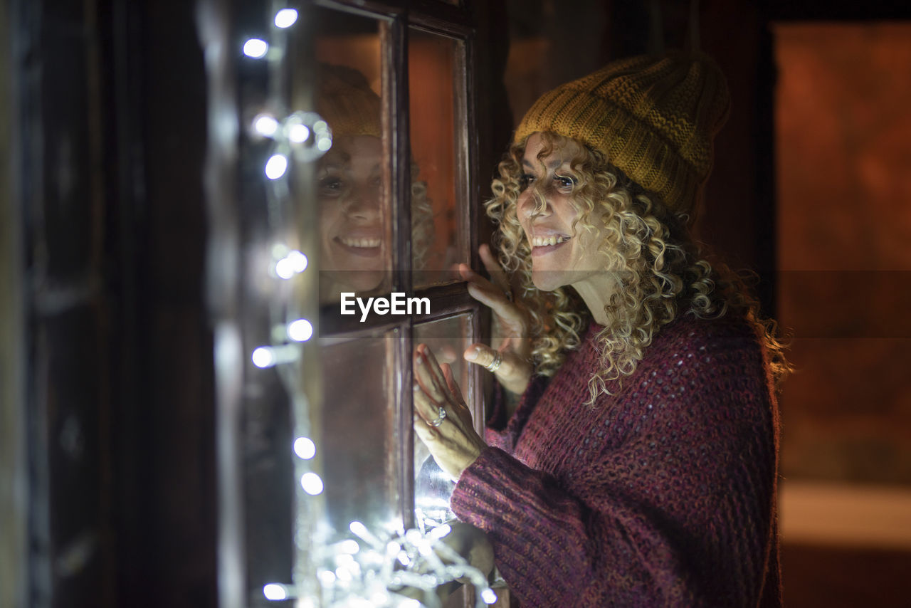 Smiling woman standing by illuminated window