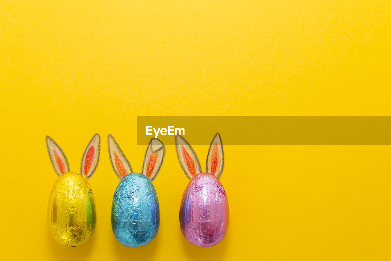 Three chocolate eggs in colorful aluminium foil with painted easter bunny ears on yellow background