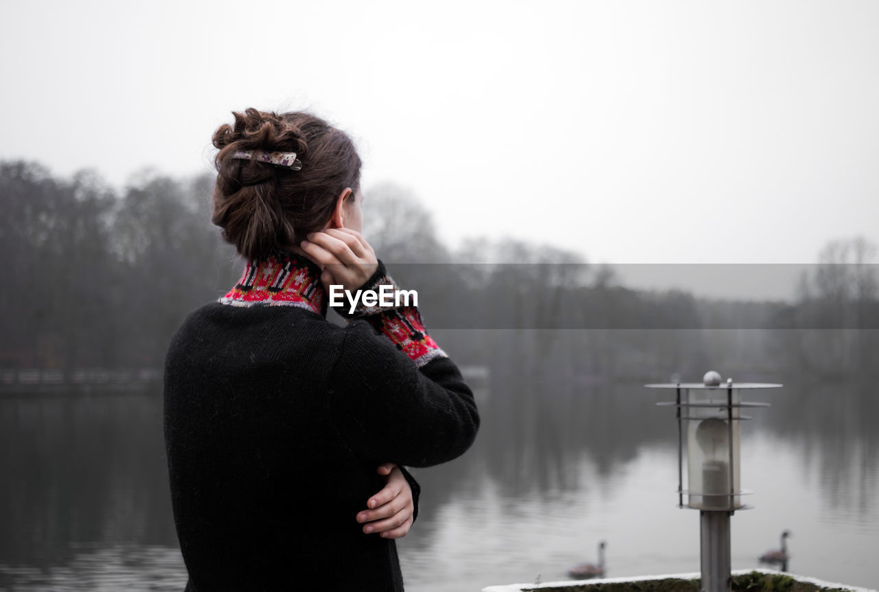A young woman stands pensively on the terrace on a cold day and looks out over the grey lake