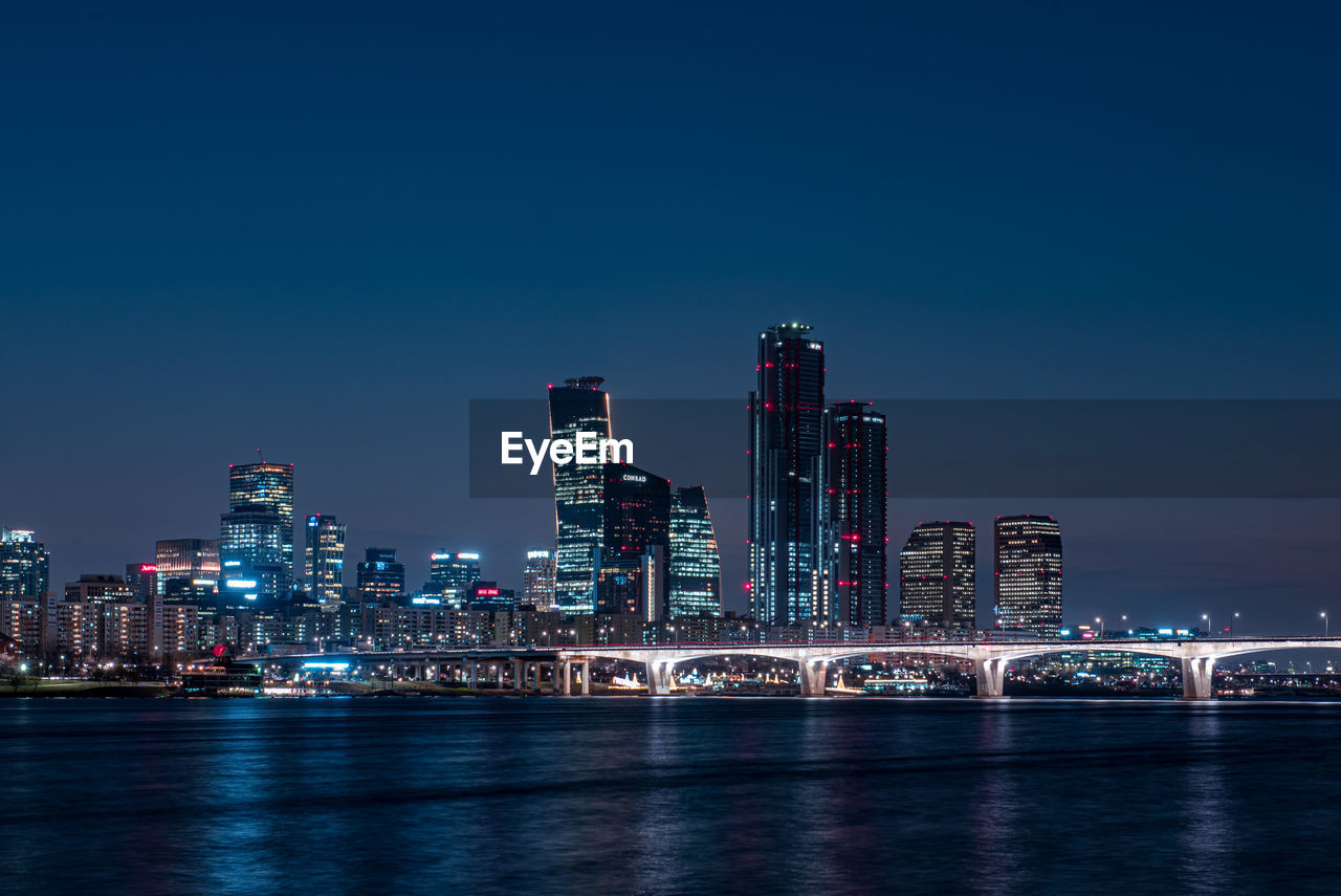 Illuminated buildings by river against sky in city at night