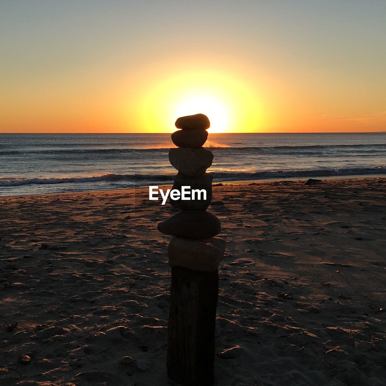 Back lit stones stack on wooden post at beach against sky during sunset
