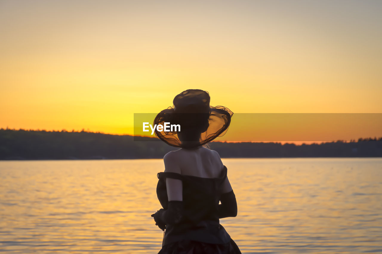 REAR VIEW OF WOMAN STANDING ON SHORE AGAINST ORANGE SKY