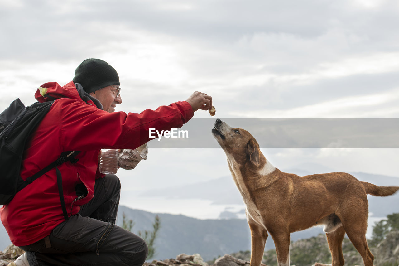Man feeding biscuit to dog on hill against sky