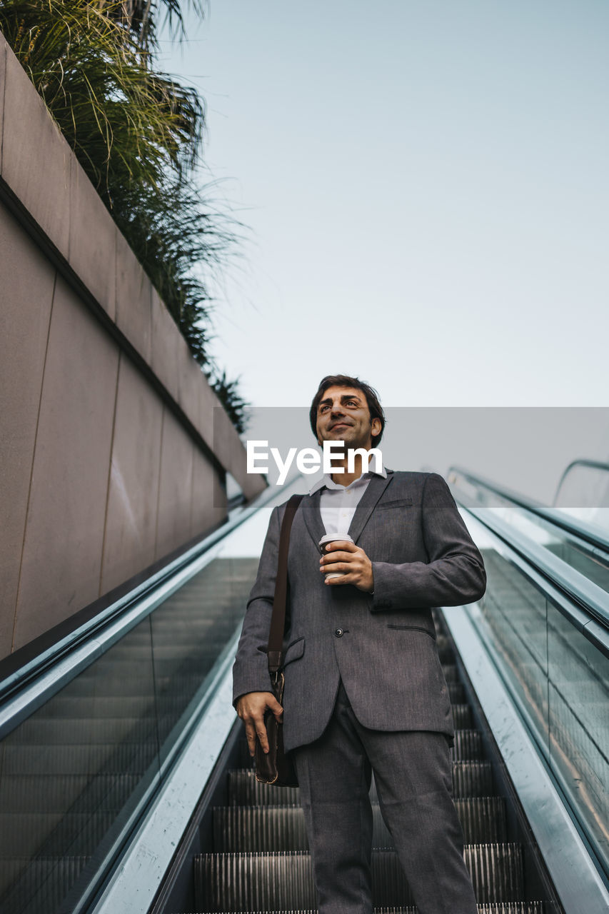 Low angle view of businessman holding coffee standing on escalator smiling against sky