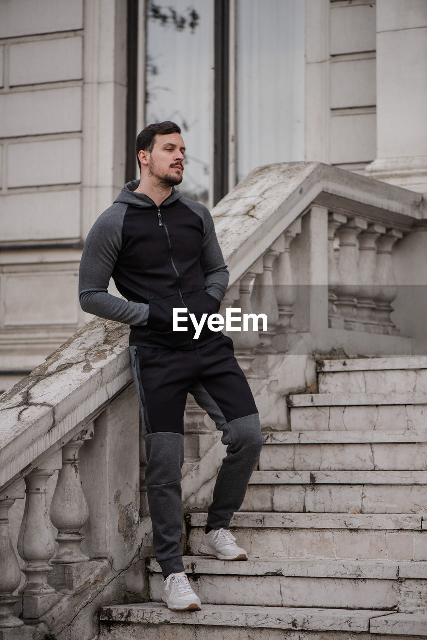 YOUNG MAN LOOKING AWAY WHILE STANDING ON STAIRCASE OF BUILDING