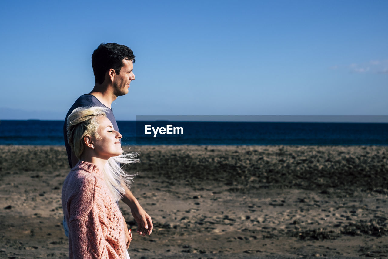 Side view of couple standing at beach against blue sky