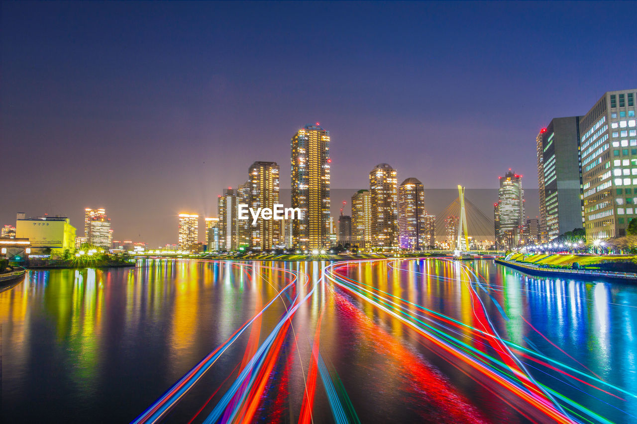 Light trails over river by illuminated buildings at night