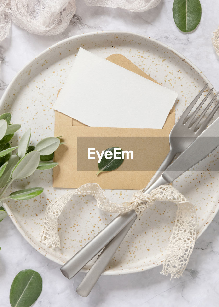 Blank paper card in envelope laying on white plate on marble table with eucalyptus branches