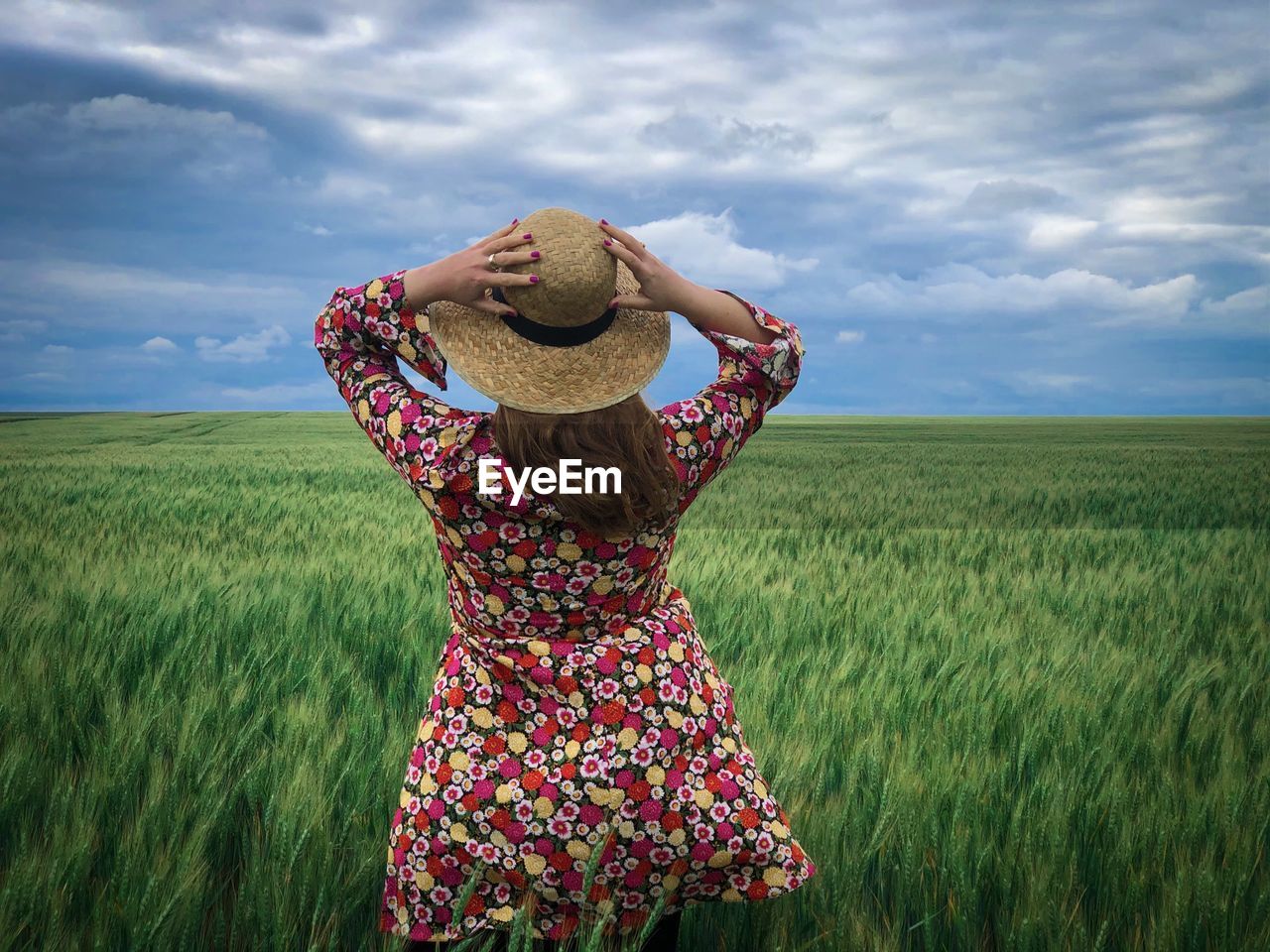 Rear view of young woman with a hat standing in a wheat field on a day with clouds