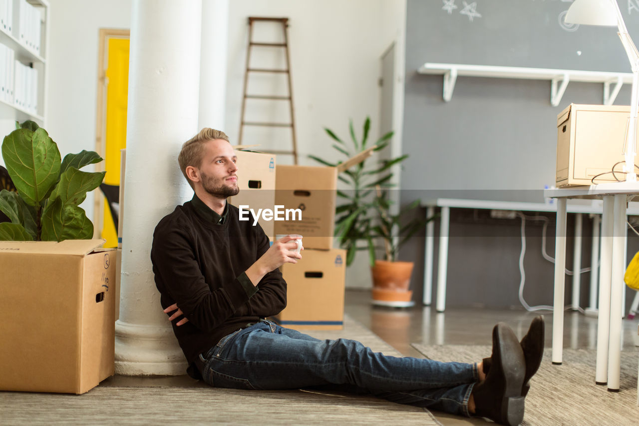 Full length of thoughtful young businessman with coffee cup sitting on floor at creative office