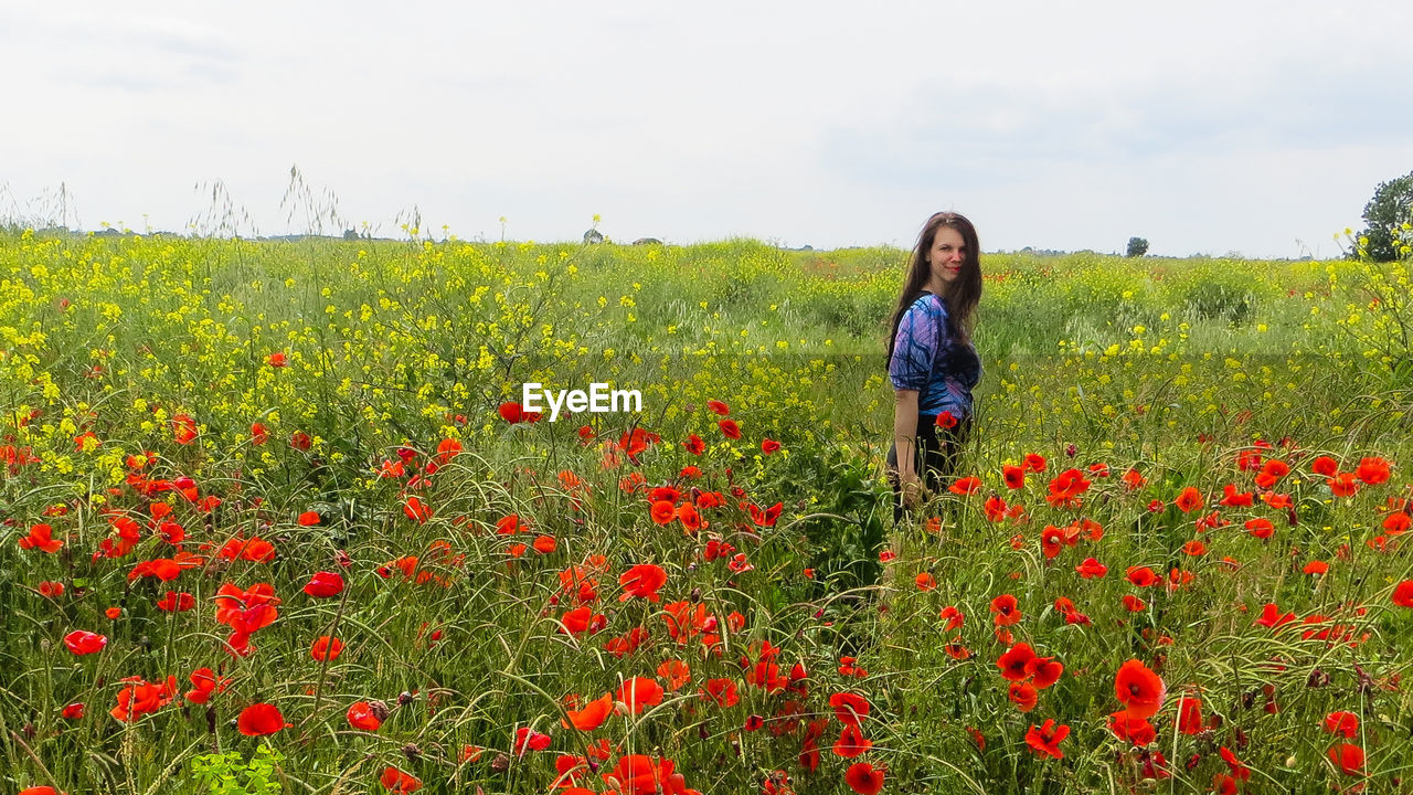 Portrait of woman standing by poppy flowers on field against sky