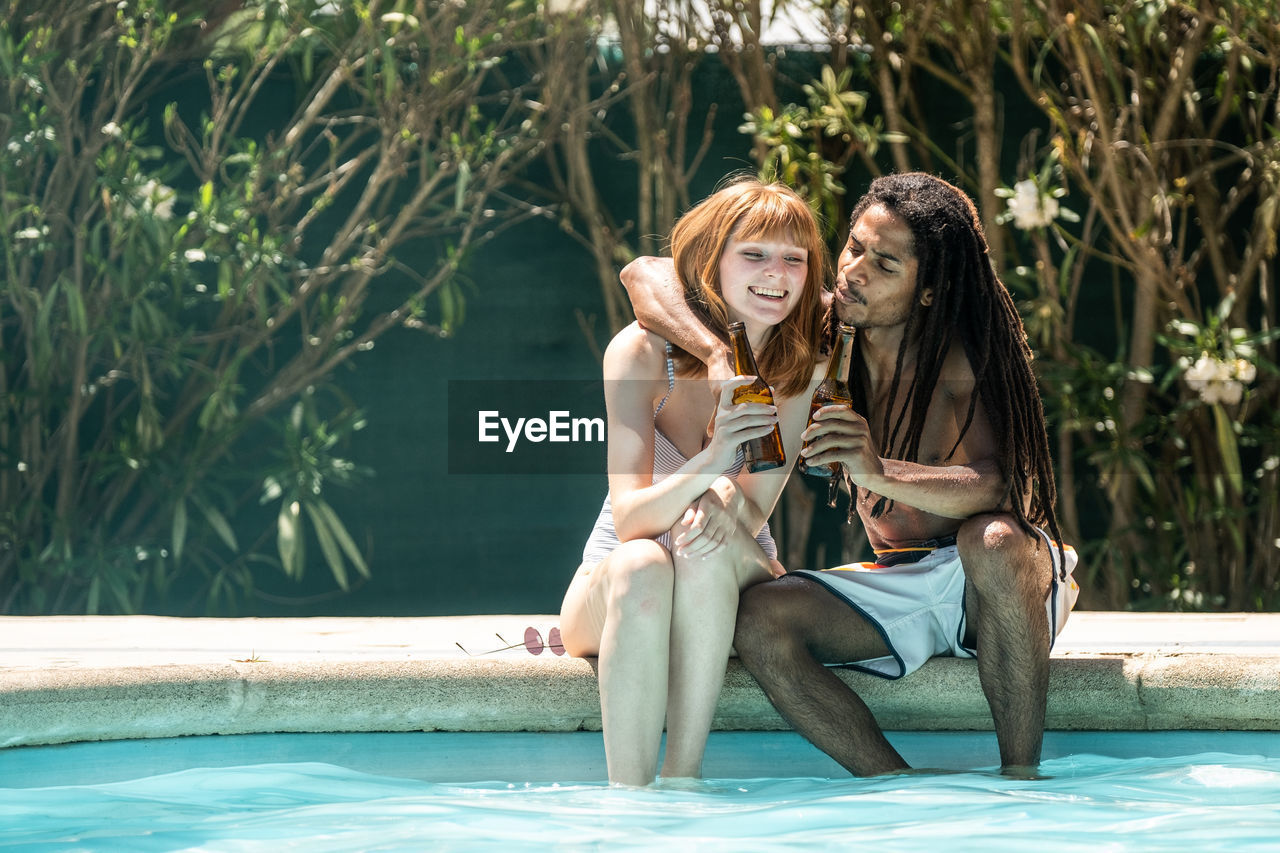 Couple toasting beer bottles while sitting at poolside