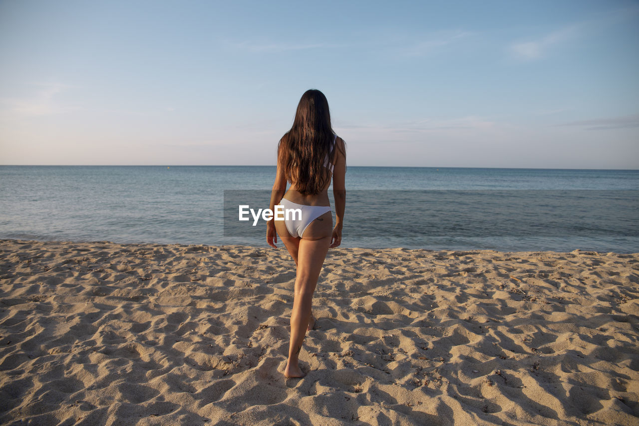 Back view of unrecognizable female in swimwear walking on sandy shore towards ocean under cloudy blue sky