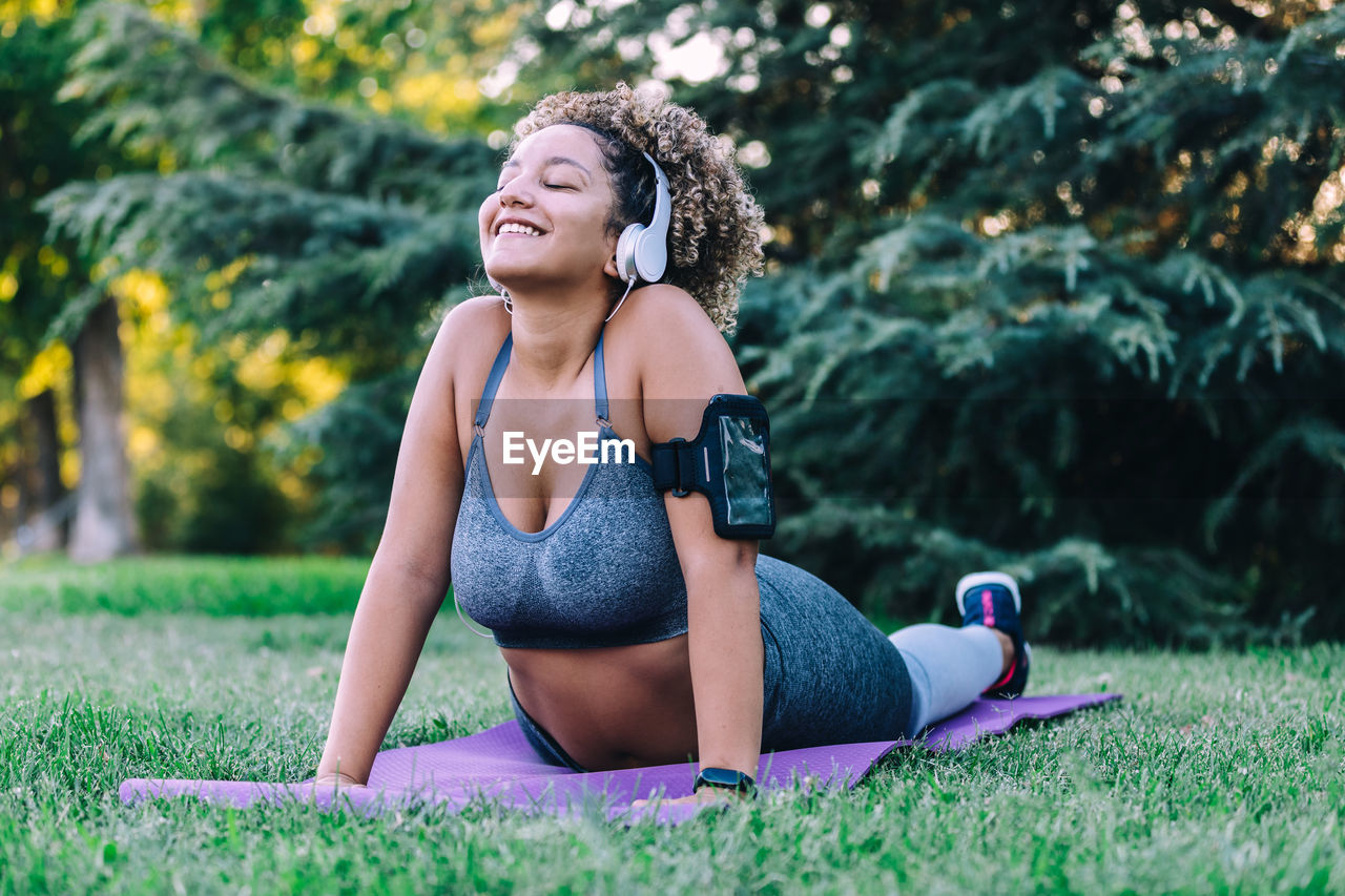 Optimistic young ethnic female in activewear listening to music with smartphone and headphones and doing back bend exercise during fitness training on green grass in park