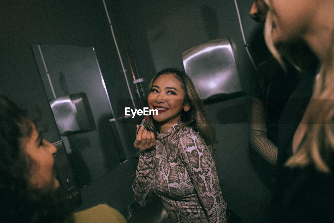 Smiling young woman applying lipstick while looking at friends in nightclub bathroom