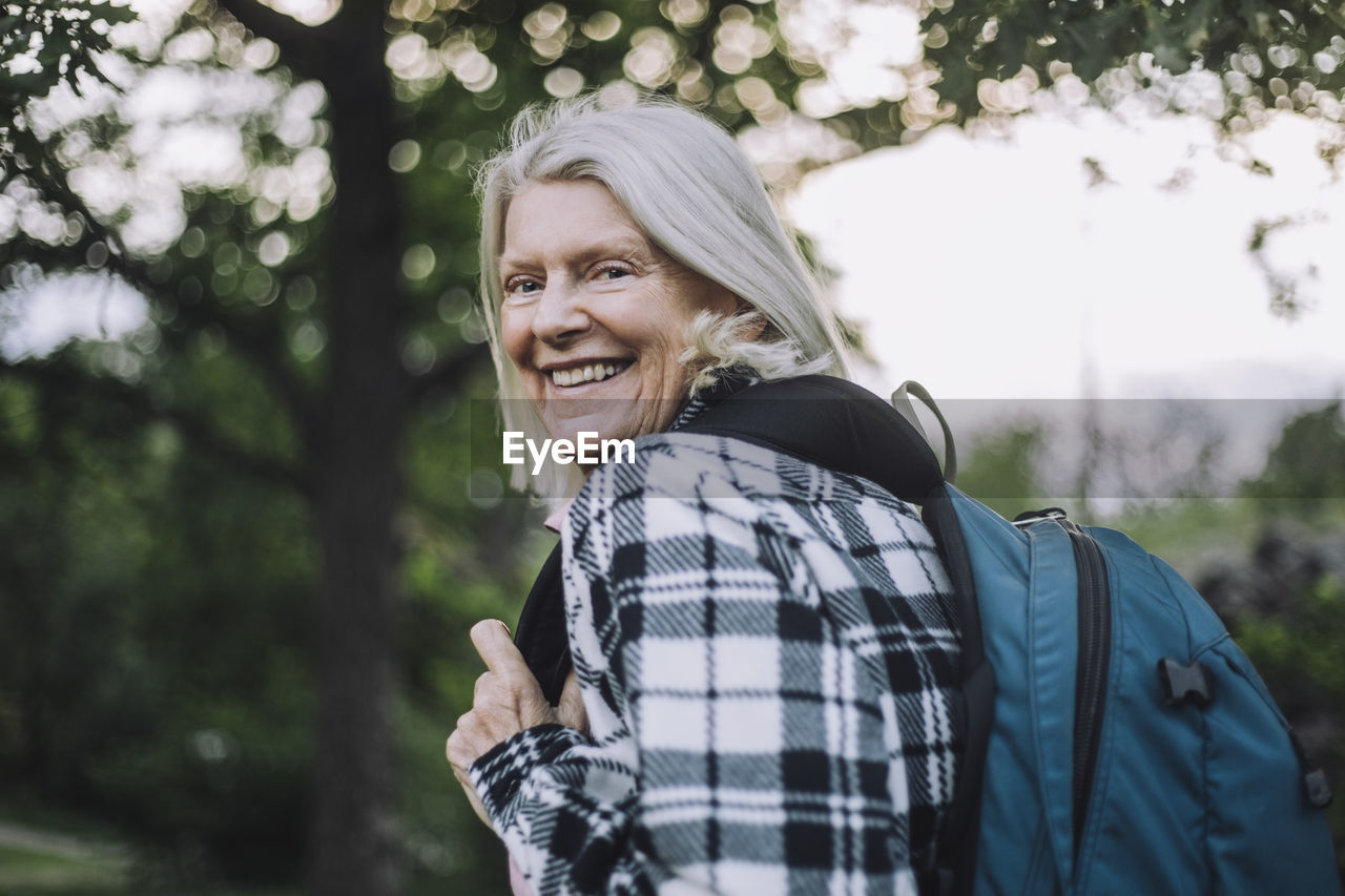 Portrait of happy senior woman looking over shoulder carrying backpack while hiking