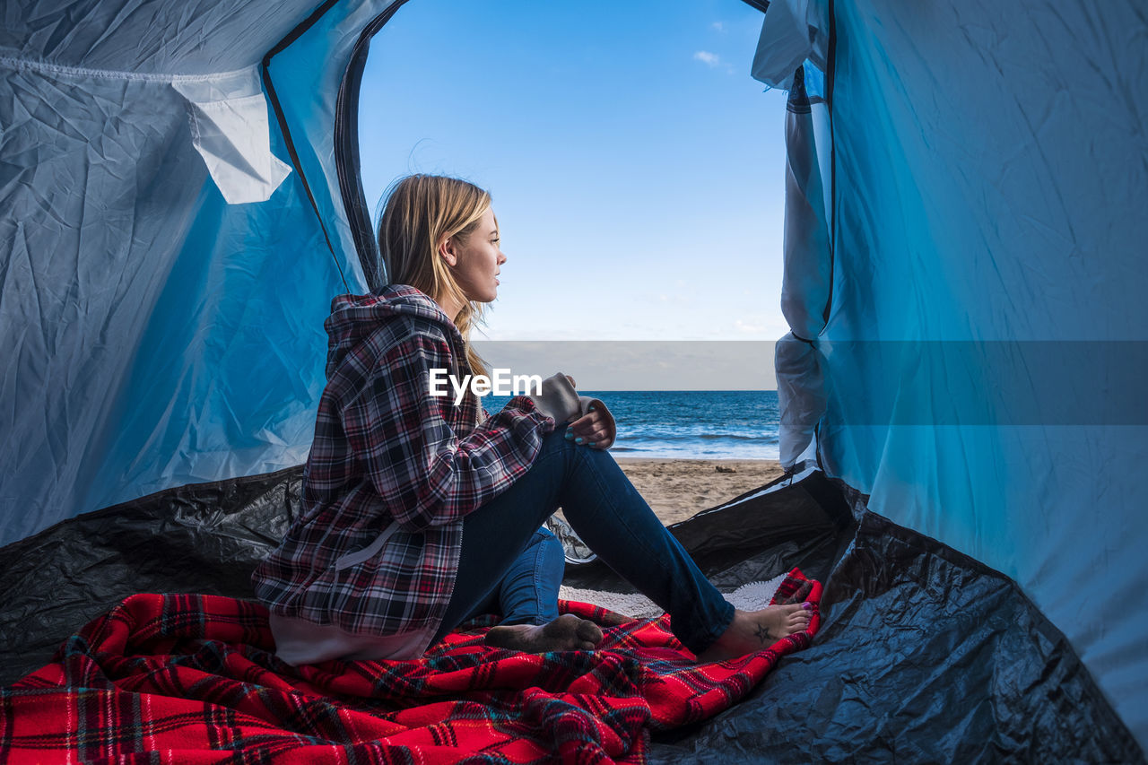 Woman sitting in tent at beach against sky