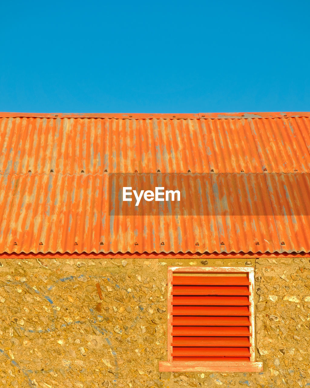 Close up of an old barn with corrugated iron roof and red shutters beneath clear blue sky