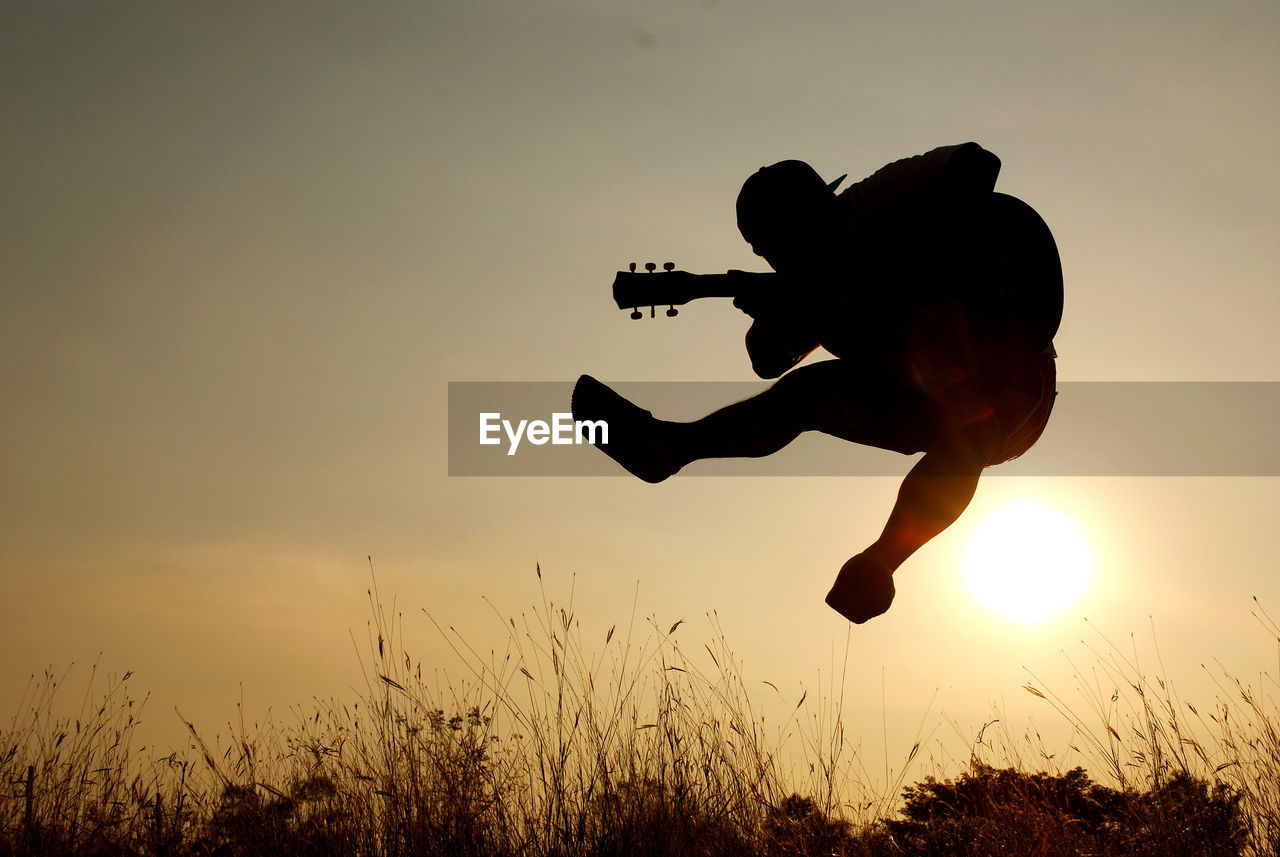 Silhouette man playing guitar while jumping against sky during sunset