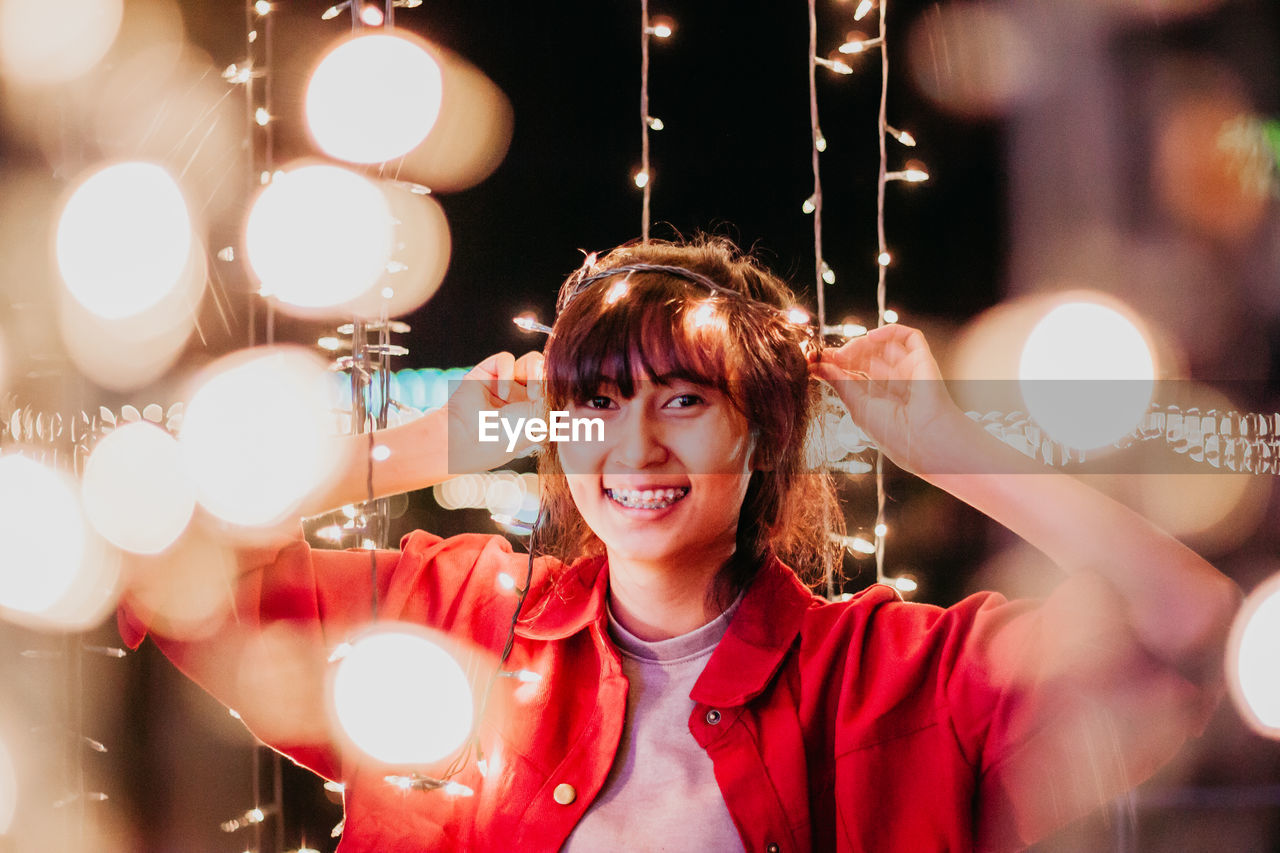 PORTRAIT OF SMILING YOUNG WOMAN WITH LIGHTS IN BACKGROUND