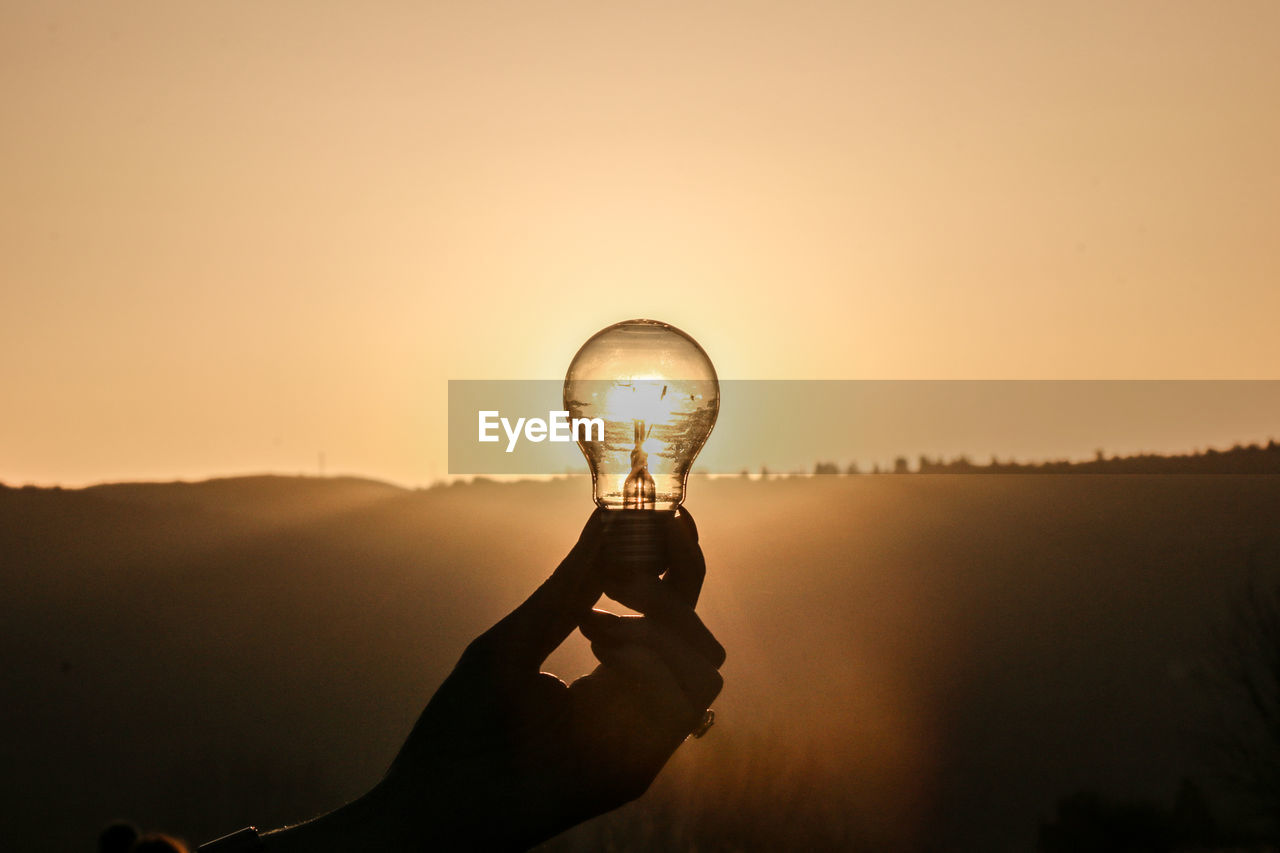 Close-up of hand holding light bulb against sky during sunset