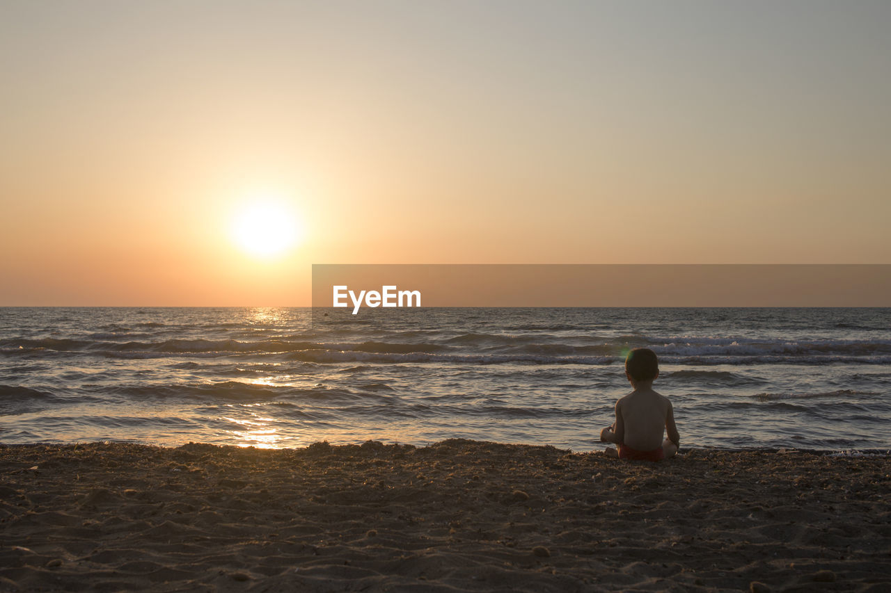 Rear view of boy sitting on beach against sky during sunset