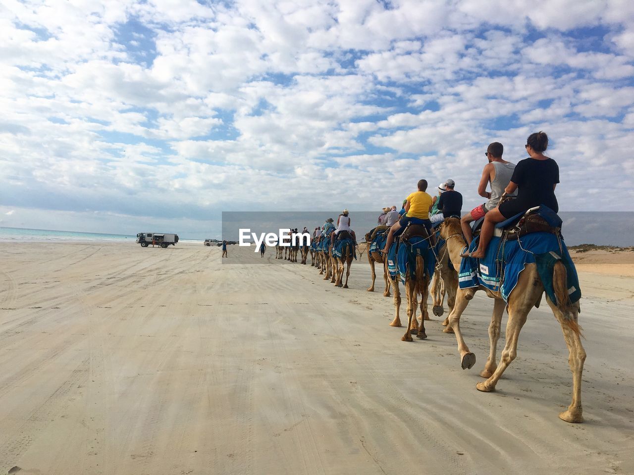 People riding on camels at beach against cloudy sky