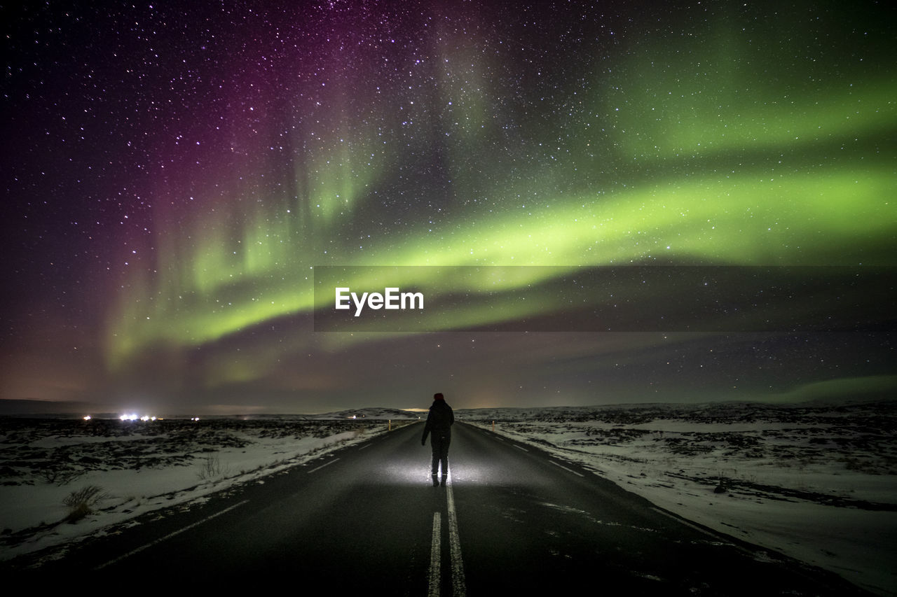 Silhouette of unrecognizable traveler standing on asphalt roadway enjoying view of green polar lights glowing in night starry sky in iceland