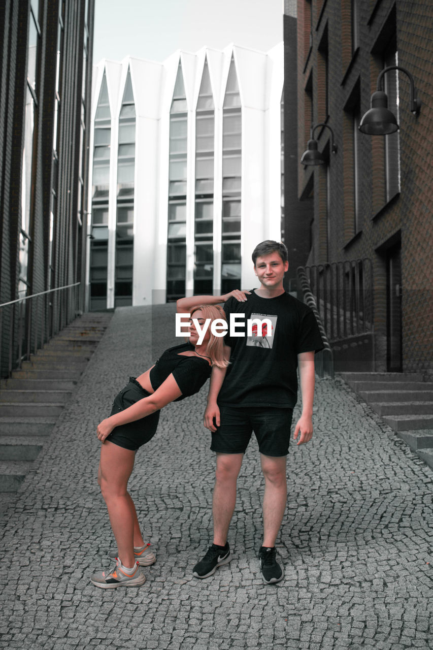 FULL LENGTH PORTRAIT OF HAPPY FRIENDS STANDING AGAINST BUILDINGS IN CITY