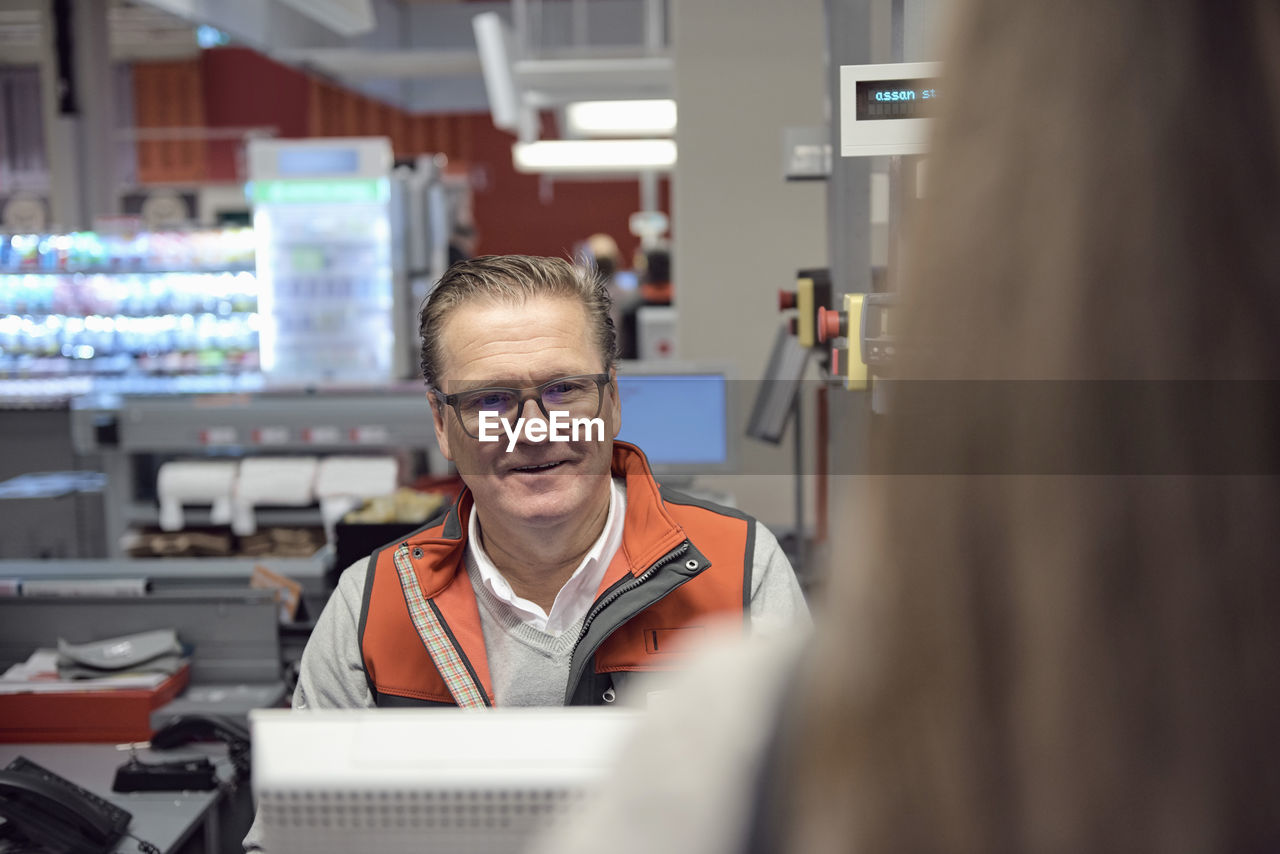 Smiling mature cashier looking at woman at checkout in supermarket