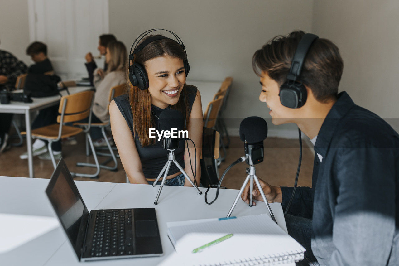 Smiling female and male students looking at each other during podcasting in classroom