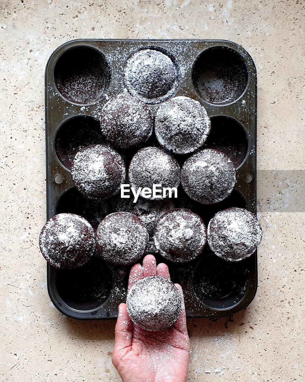 Directly above shot of hand holding cupcake over baking tray on stone table