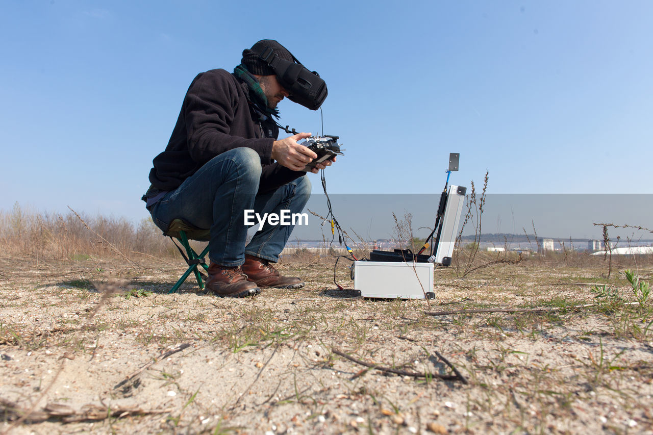 Man operating drone with remote control while sitting on field against sky