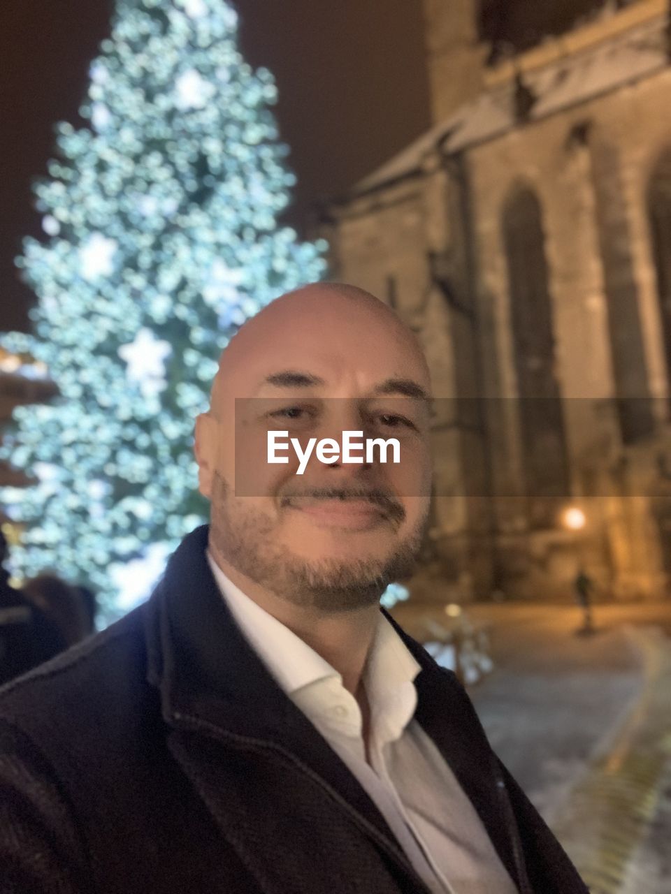 one person, men, adult, christmas tree, portrait, architecture, city, building exterior, smiling, person, tree, business, christmas, happiness, headshot, celebration, winter, clothing, formal wear, focus on foreground, businessman, night, standing, built structure, emotion, beard, waist up, holiday, nature, front view, looking at camera, city life, plant, looking, young adult, street, outdoors