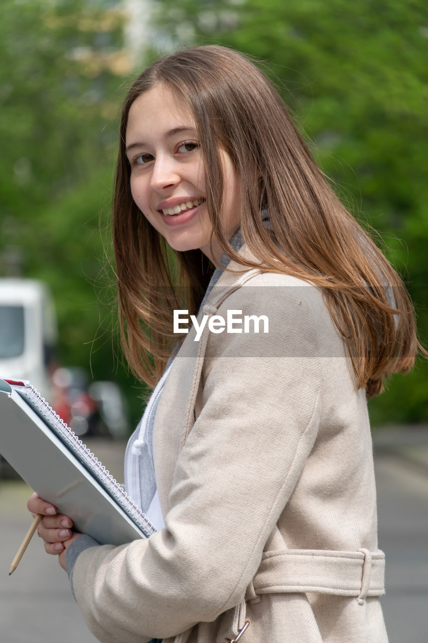 Portrait of smiling teenage girl holding books against trees