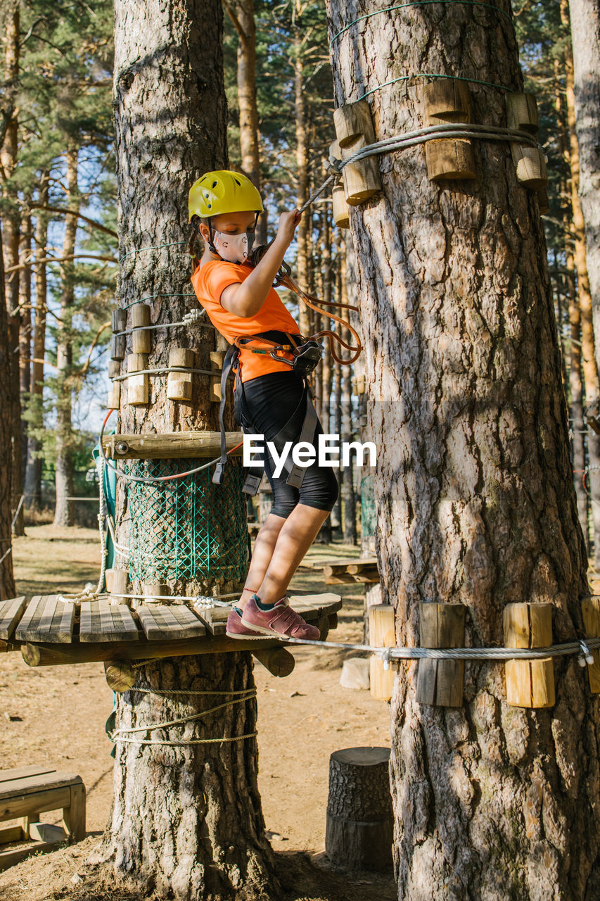 Concentrated kid in medical mask and safety equipment standing on wooden suspension bridge in adventure park during covid 19 epidemic