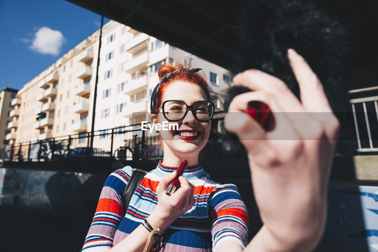 Young woman looking at mobile phone while applying lipstick in city
