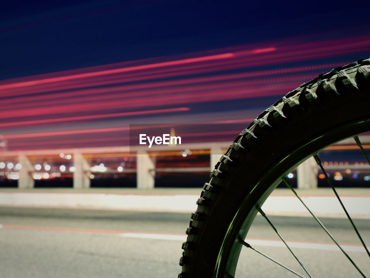 Close-up of bicycle tire by illuminated light trails on bridge at night