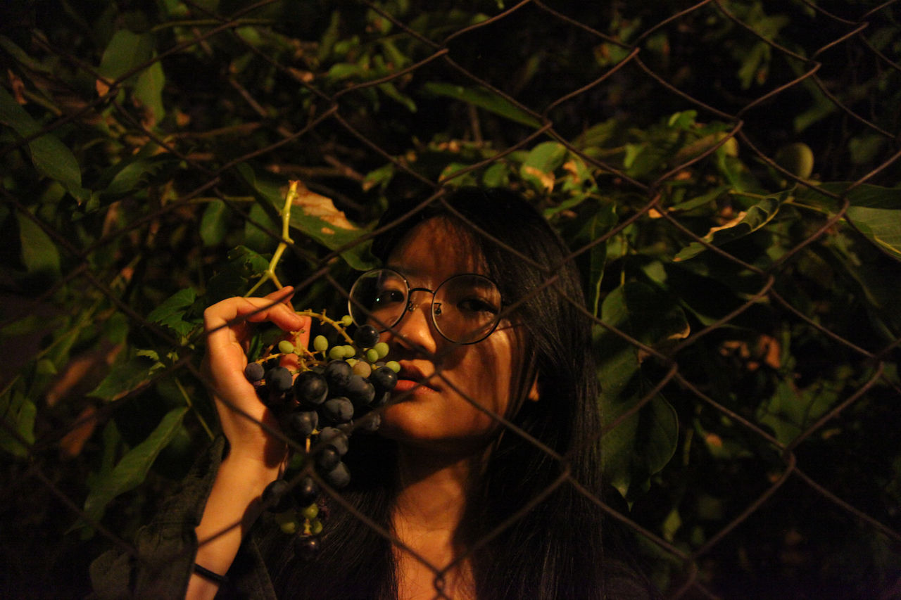 Portrait of woman holding grapes while standing against chainlink fence