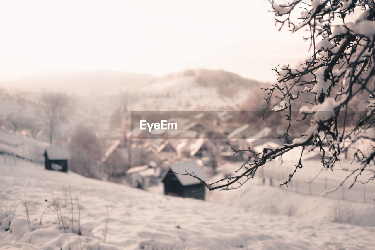 Scenic view of village in front of snowcapped mountains against sky. branches  in  foreground.