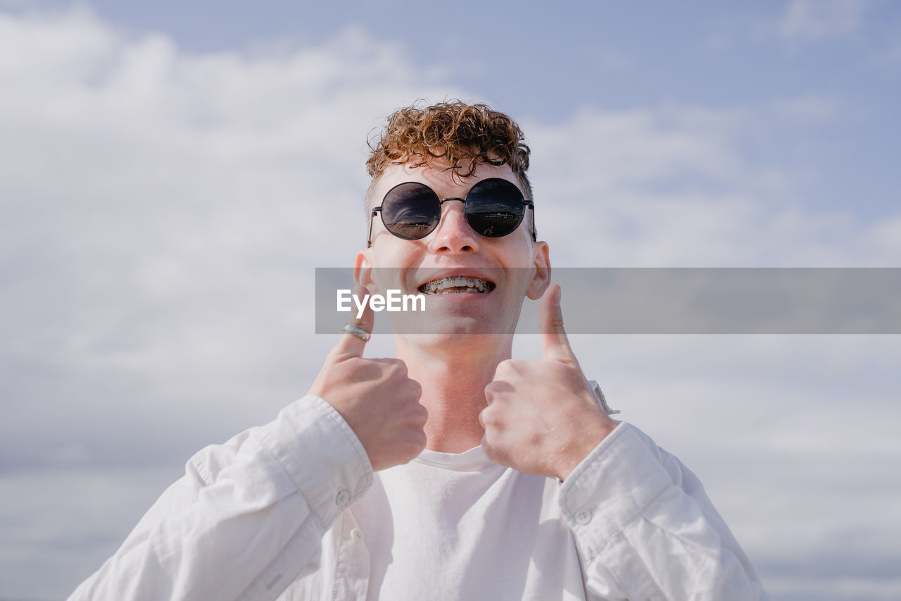 A young guy stands against the background of the sky shows thumbs up, smiles a wide smile with brace