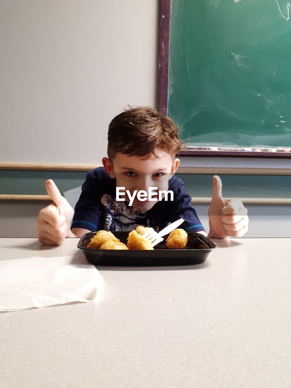 Portrait of happy boy showing thumbs up while sitting with food on table in classroom