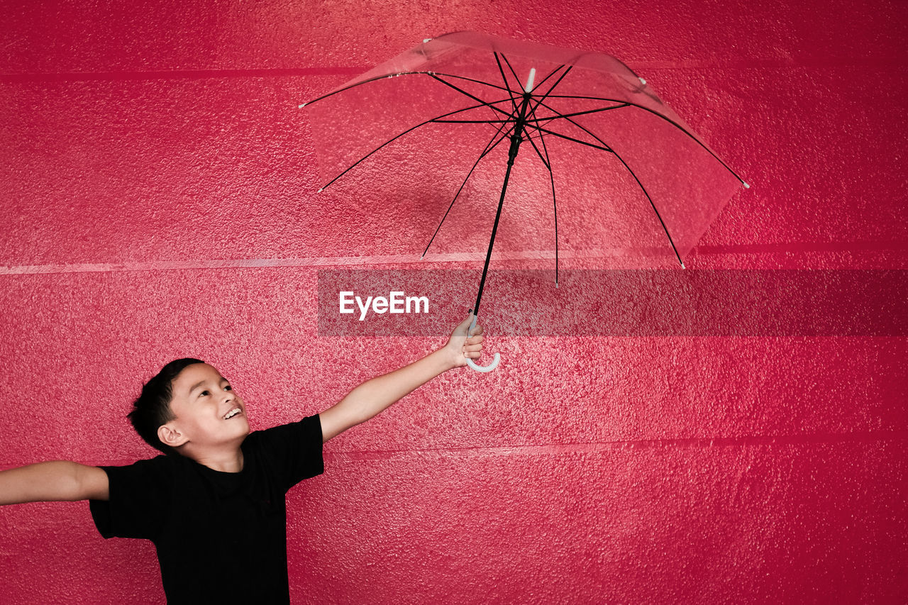 Boy holding umbrella while standing against red wall